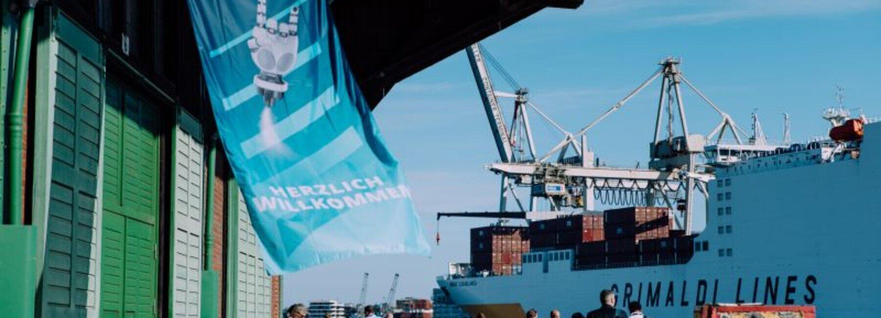 Blick auf ein Hafengebäude mit Personen und einem Containerschiff