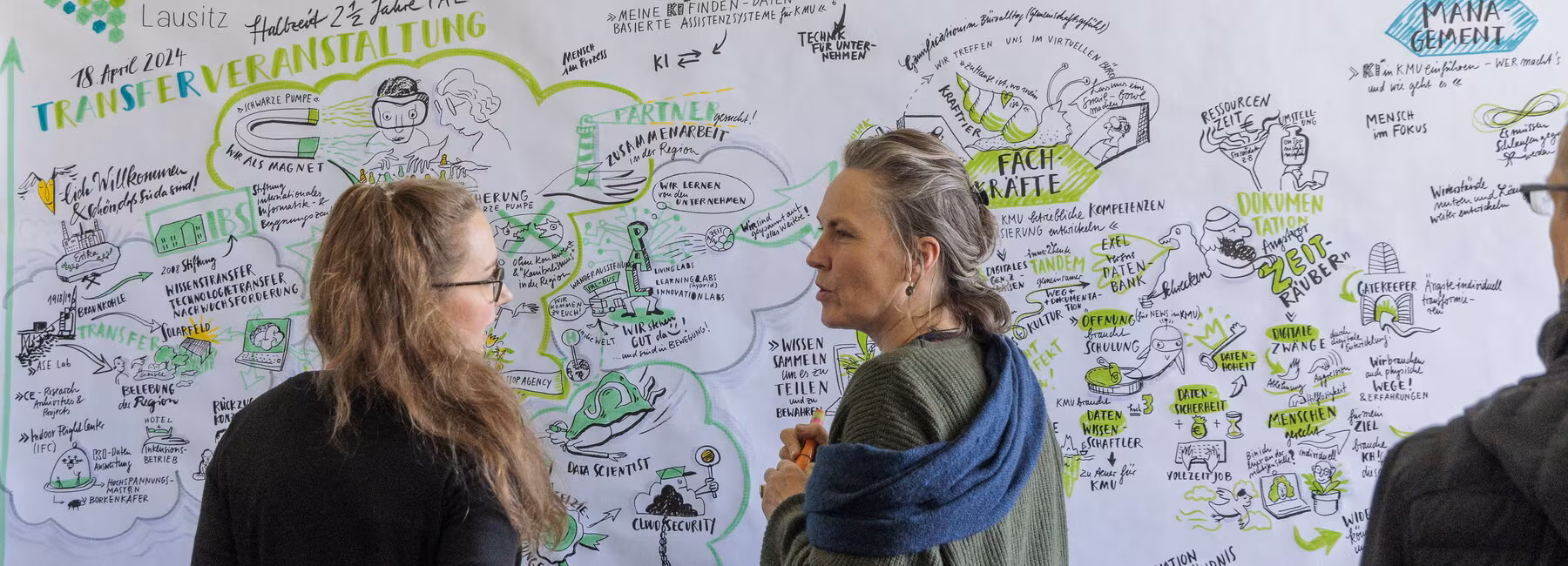 Zwei weibliche Personen stehen vor einem Banner mit grafischen Elementen