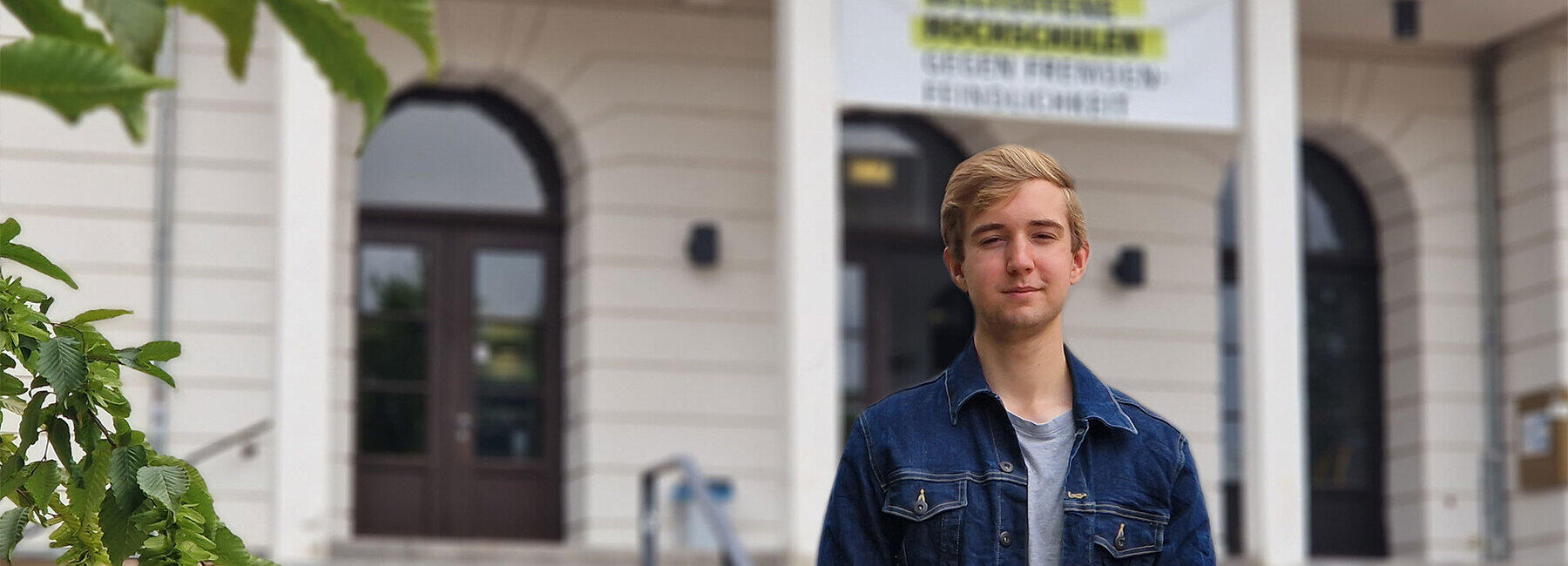 Ein junger Mann steht vor einem Haus mit einem Transparent "weltoffene Hochschule".