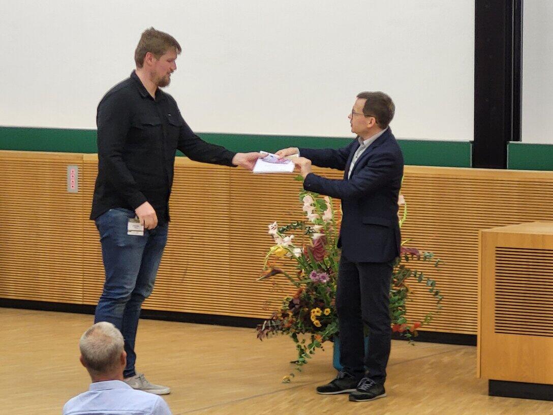 Zwei männliche Personen stehen auf einer Bühne, ein Preis wird übergeben