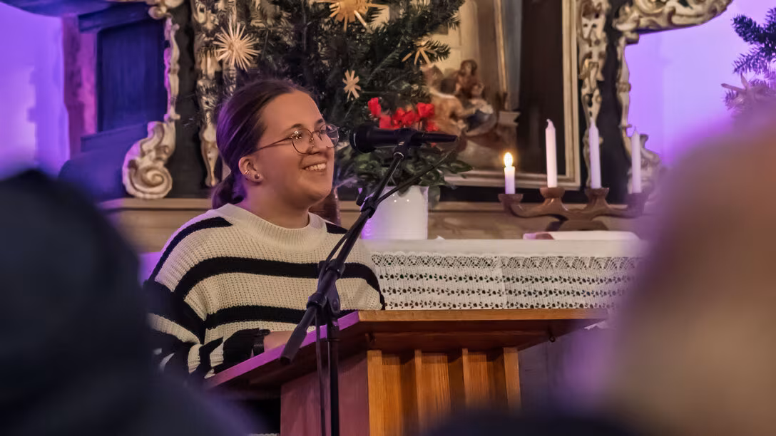 Eine junge weibliche Person steht lächelnd am Pult vor dem Altar uns spricht in ein Mikrofon.