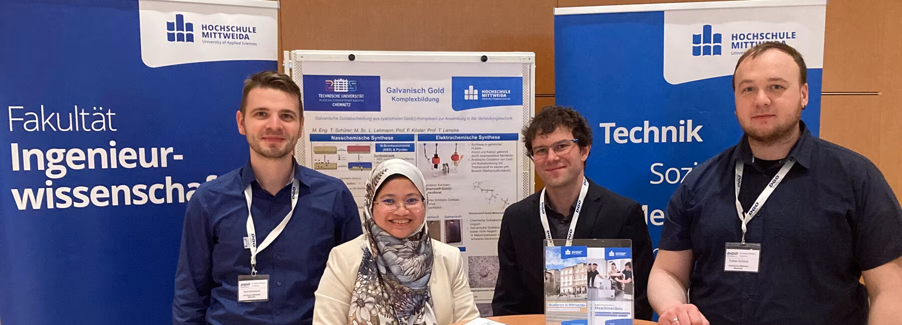 vier Personen stehen um einen Tisch, im Hintergrund Rollups und ein Poster der Hochschule Mittweida
