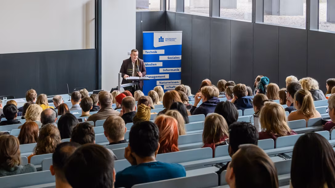 Blick von hinten in einen besetzten Hörsaal. Unten im Hörsaal im Bildhintergrund steht eine männliche Person an einem Pult, dahinter das Rollup mit dem Logo der Hochschule Mittweida und den Begriffen „Technik“, „Soziales“, „Medien“, Informatik“, Management“ und „Naturwissenschaft“.
