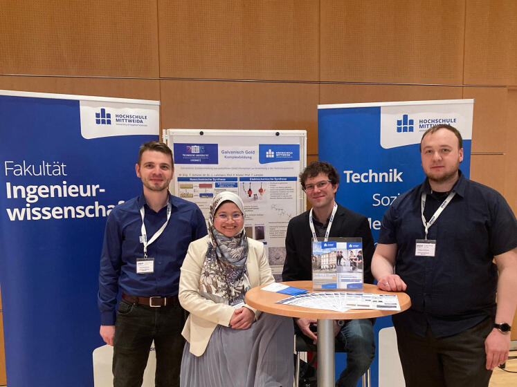 vier Personen stehen um einen Tisch, im Hintergrund Rollups und ein Poster der Hochschule Mittweida