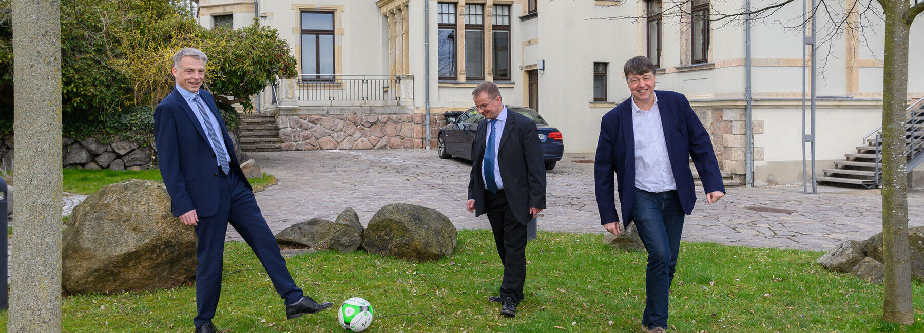 Drei männliche Personen in Anzügen spielen Fußball auf der Wiese vor einem historischen Gebäude, der Direktorenvilla der Hochschule Mittweida.
