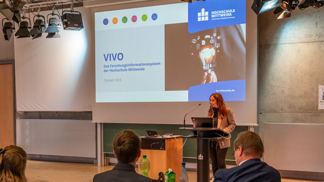 Weibliche Person steht vor einer Projektionswand und spricht in ein Mikrofon