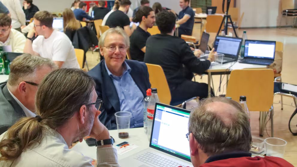 Zahlreiche Personengruppen an Tischen, auf denen Laptops und Getränke stehen. Ein einzige männliche Person in der Bildmitte schaut in die Kamera.
