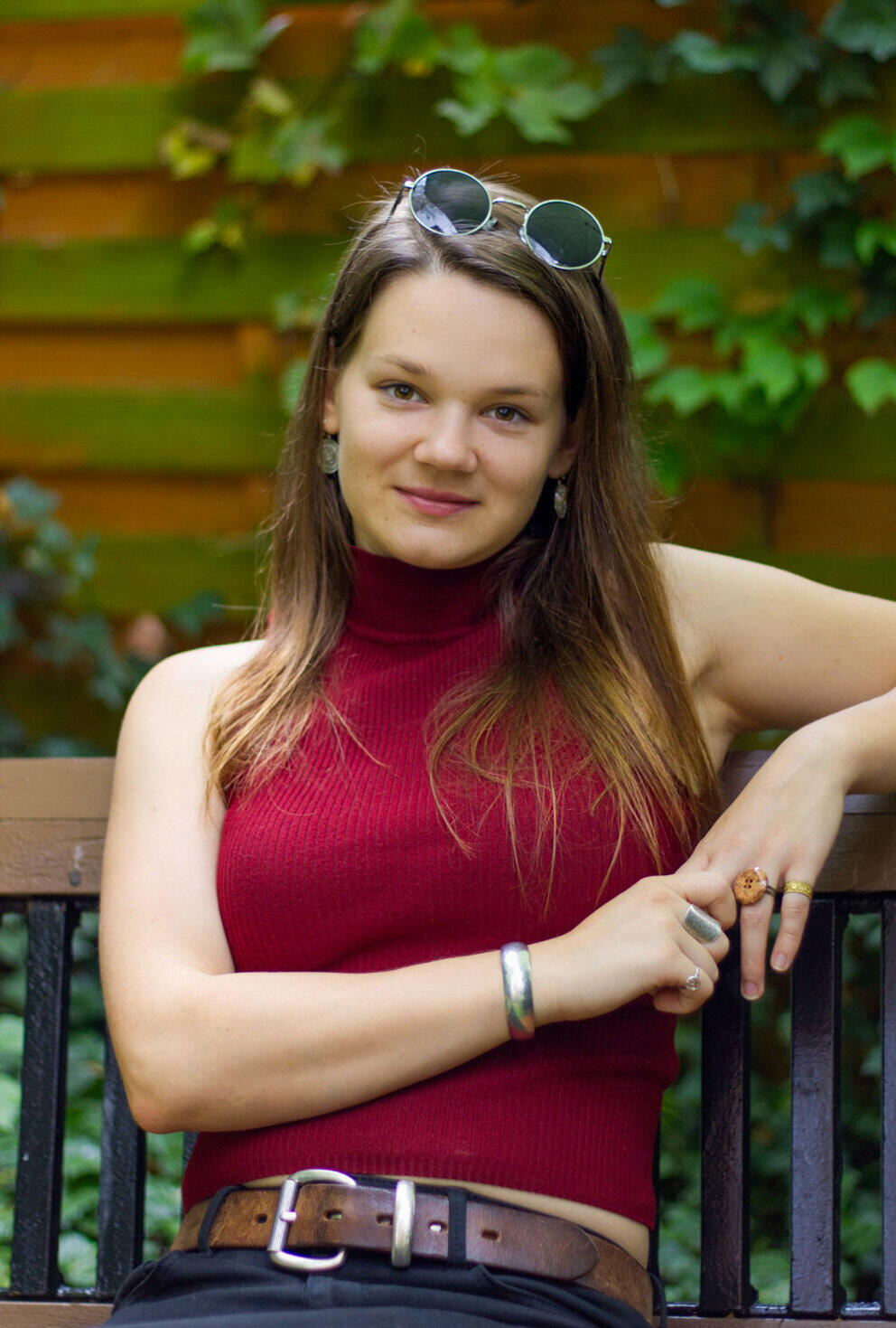 Eine junge Frau sitzt lässig auf einer Bank im Grünen, die Sonnenbrille ins Haar geschoben, und lächelt in die Kamera.