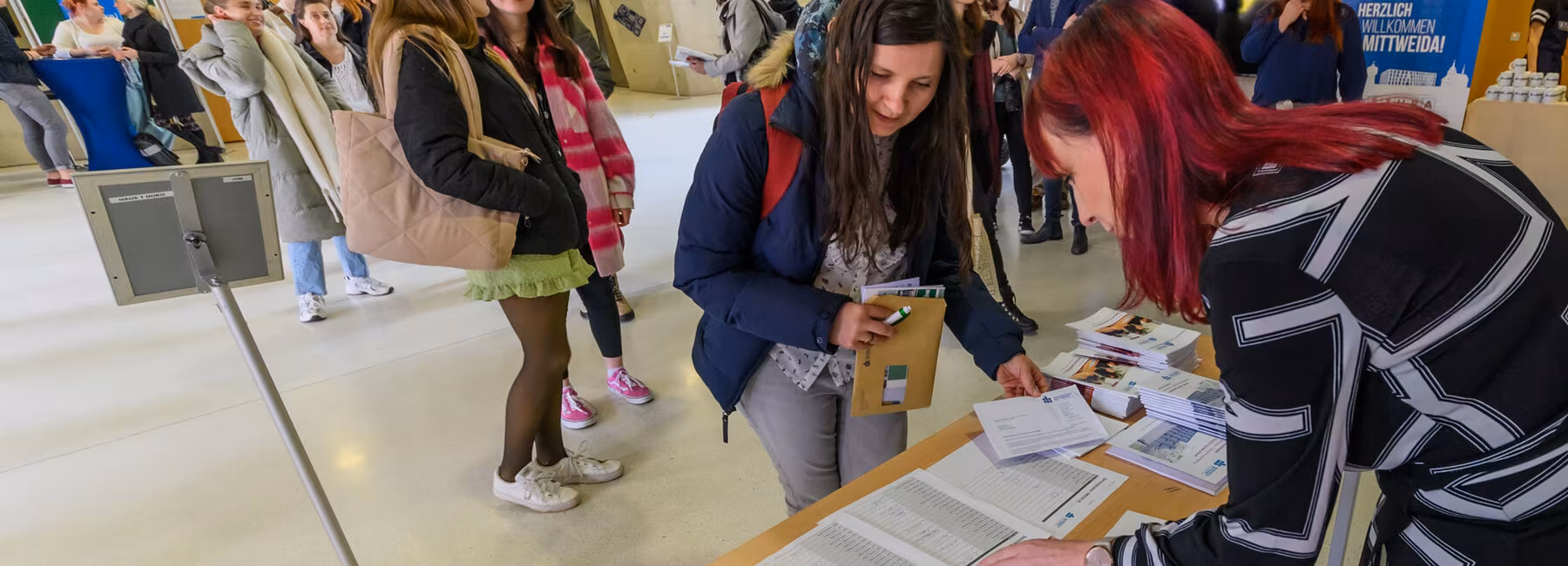Ein großes Foyer. Einige Menschen stehen in einer Schlange. Die erste Frau in der Schlange steht vor einem Tisch im Vordergrund. Ihre gegenüber auf der anderen Seite des Tisches steht eine weitere weibliche Person. Beide beugen sich über Unterlagen auf dem Tisch.