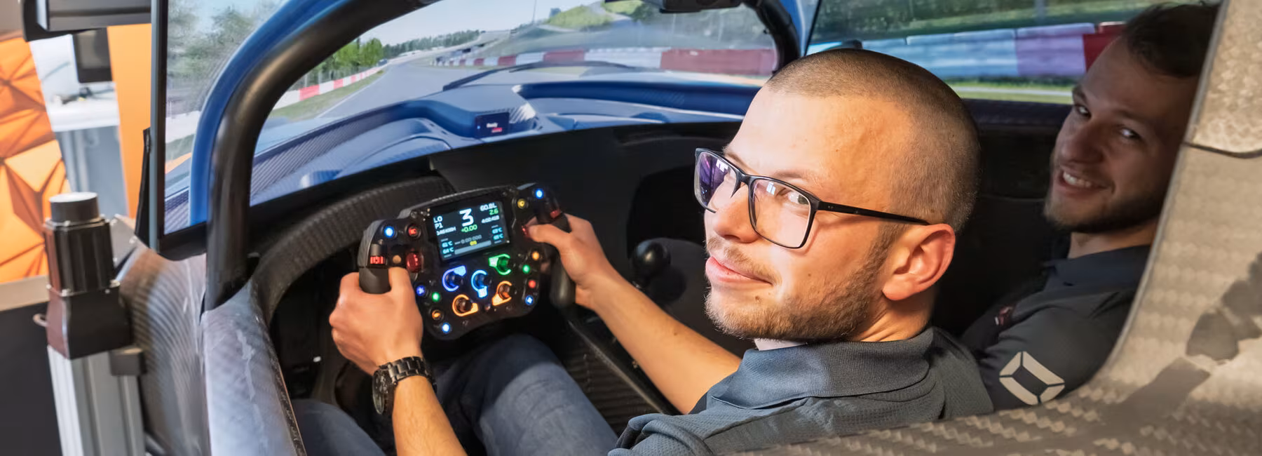 Zwei junge Männer im Cockpit eines Rennwagens, das Teil einer Simulationsplattform ist, in der das Umfeld Rennstrecke auf großen Monitoren dargestellt ist. Der linke Mann fasst mit beiden Händen das Lenkrad. Beide schauen nach hinten in Richtung Kamera.