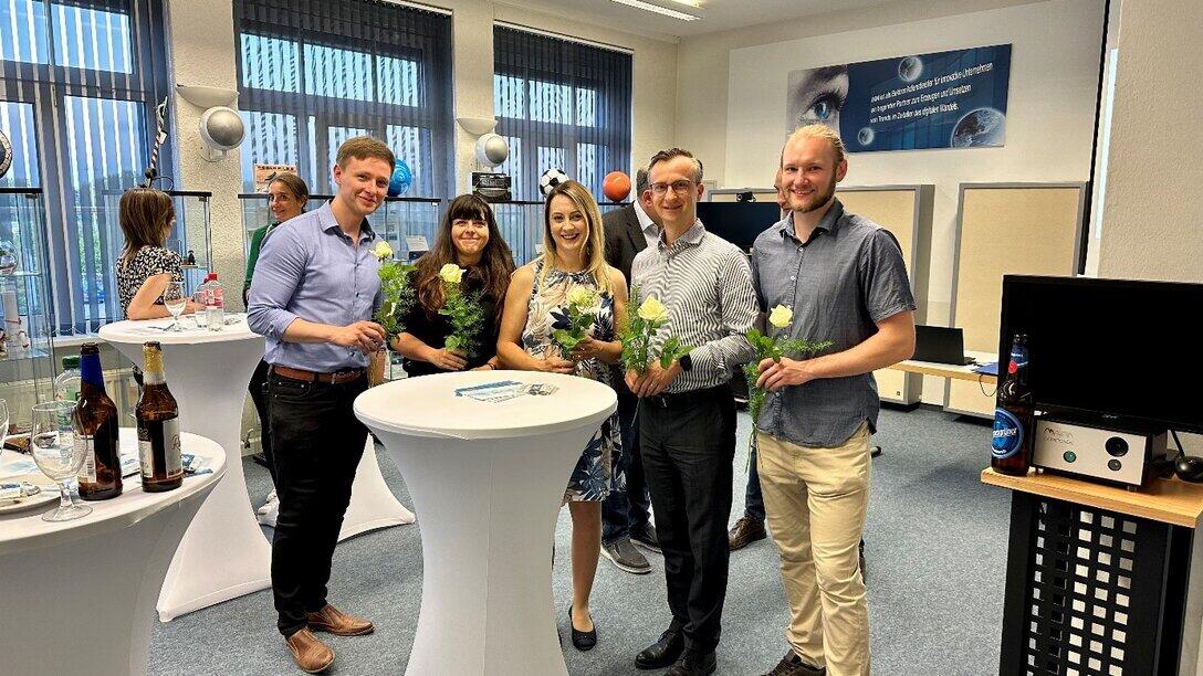 fünf Personen stehen mit einer Blume an einem Stehtisch, im Hintergrund stehen zwei weitere Personen an einem Stehtisch