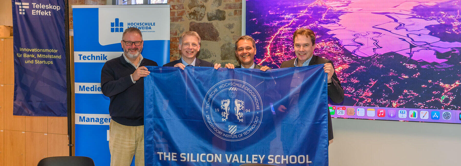 Vier männliche Personen halten freudenstrahlend eine Fahne mit der Ausfschrift "Siliccon Valley School". Im Hintergrund ist eine Luftaufnahme vom Silicon Valley in Klaifornien zu sehen, sowie drzwei Rollups von Teleskopeffekt und Hochschule Mittweida.
