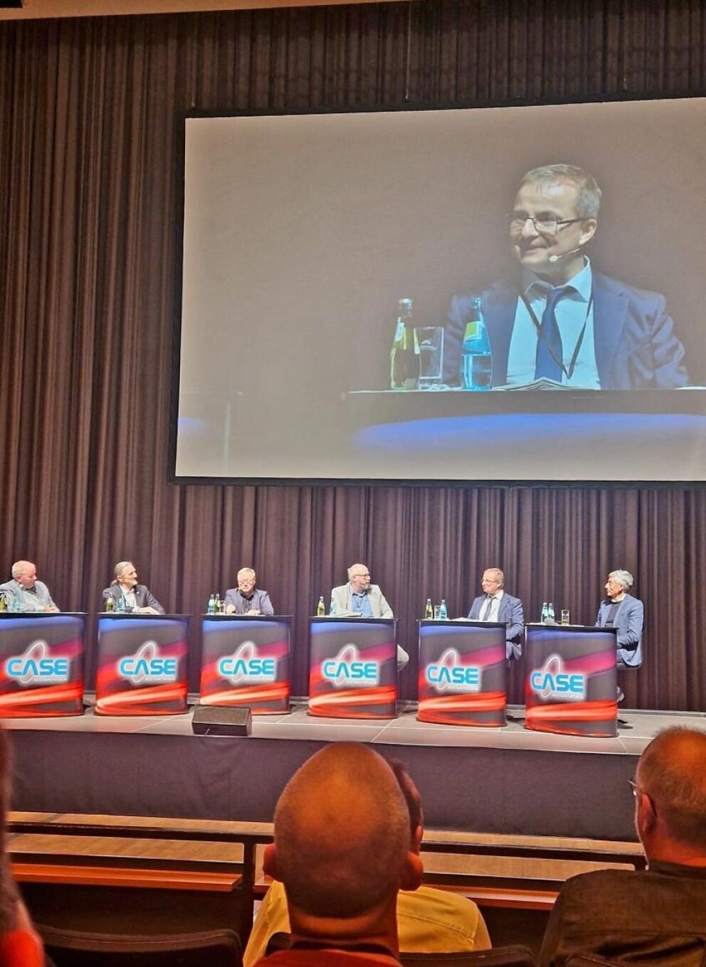 Sechs männliche Personen sitzen hinter Tischen auf einer Bühne, auf einer Präsentationswand ist eine Person im Portrait abgebildet