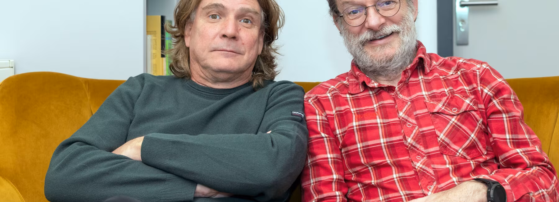 Two men are sitting on a sofa, smiling cheerfully at the camera.
