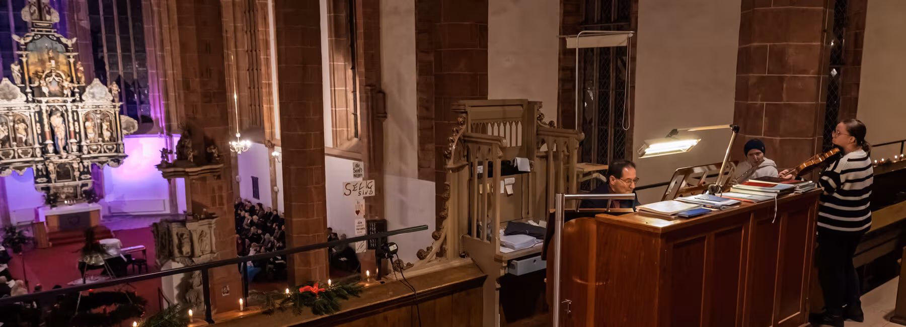 View of the gallery and the nave. In the gallery, a man is sitting at the organ console; to his right stand two women, one playing the violin and the other the flute.