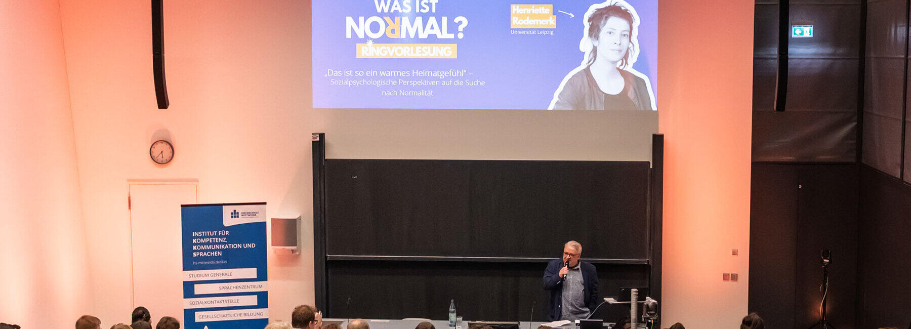 Blick von hinten über die Köpfe der Besucher:innen hinweg in den Hörsaal. Hinter dem Pult spricht eine männliche Person mit Mikrofon in der Hand zum Publikum. Links steht ein blaues Rollup u.a. mit dem Text „Institut für Kompetenz, Kommunikation und Sprachen“. Das Beamerbild an der Wand zeigt die Begrüßungsfolie u.a. mit dem Text „Was ist normal? Ringvorlesung“ und dem Titel der ersten Vorlesung: „Das ist so ein warmes Heimatgefühl“ – Sozialpsychologische Perspektiven auf die Suche nach Normalität“. Daneben ist die Referentin Henriette Rodemerk abgebildet.