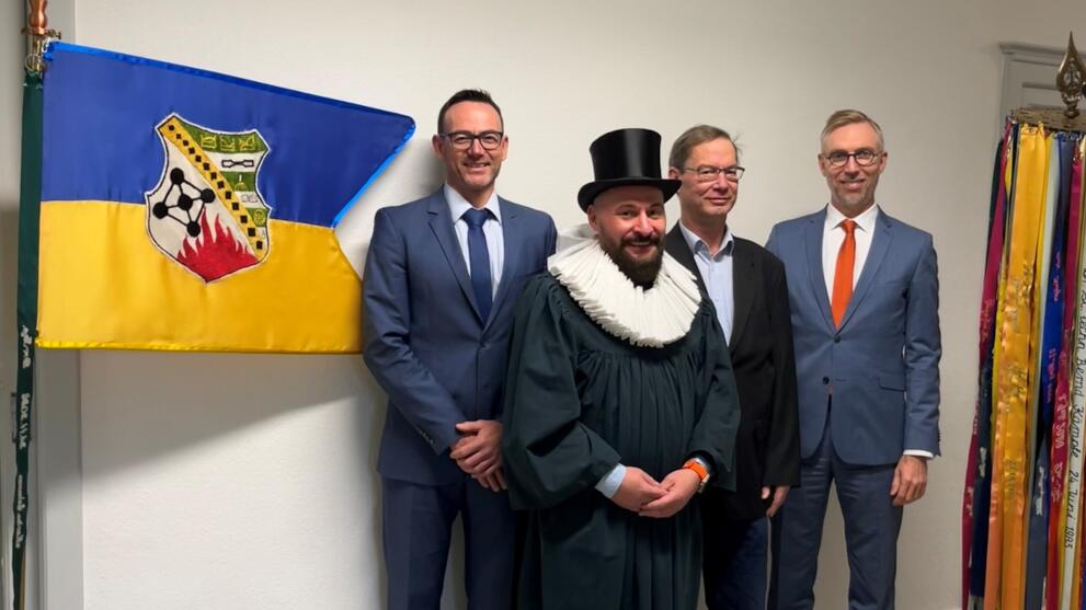 Vier männliche Personen vor einer mit einer Fahne geschmückten Wand