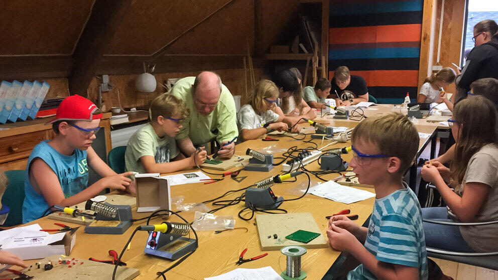 Elf Kinder mit Schutzbrillen sitzen an einem großen Tisch, auf dem sich kleinen Lötstationen und elektronische Bausätze befinden. Eine erwachsene Person beugt sich zu einem Jungen und unterstützt ihn beim Löten.