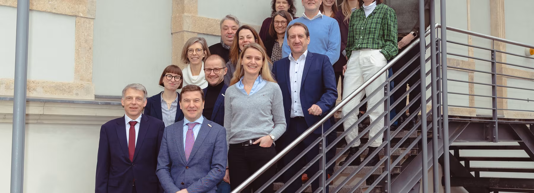 Auf den Stufen einer stählernen Außentreppe zu einem Gebäude stehen 17 lächelnde Personen.