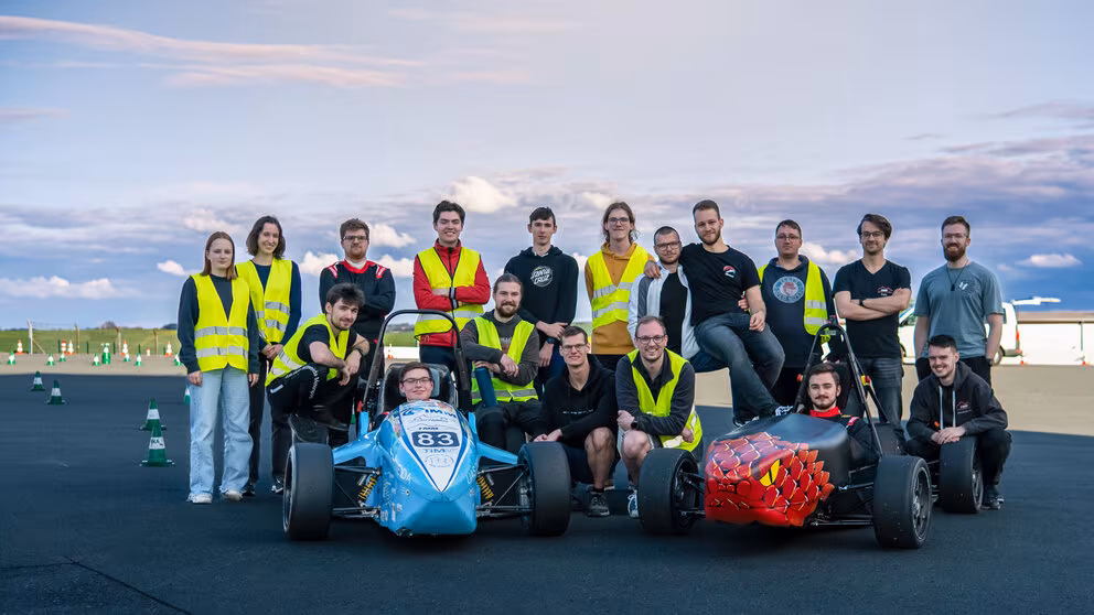 Gruppenfoto mit 18 jungen Personen, teilweise in gelben Warnwesten, und einem blauen und einem roten offenen Rennwagen auf einer geteerten Fläche. Zwei der Personen sitzen in den Rennwagen, fünf knien links und rechts davon, die anderen stehen dahinter. Im Bildhintergrund sind rot-weiße Pylone aufgestellt.