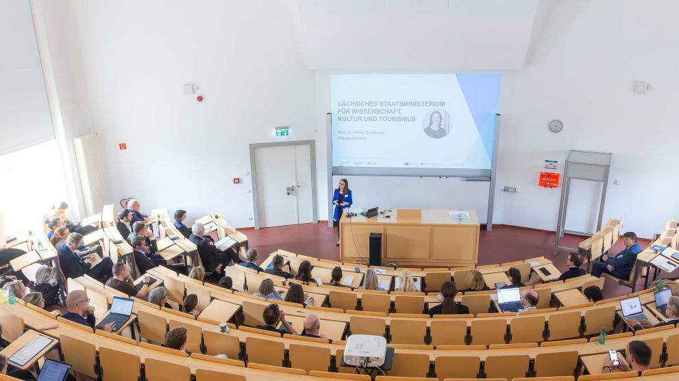 A perspective view of a lecture theatre; some seats are occupied; a woman in a blue blazer is standing at the lectern