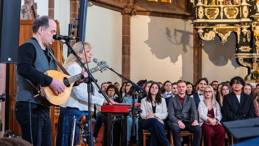 Musikduo seitlich im Vordergrund. Dahinter sitzen Absovent:innen auf Stühlen.