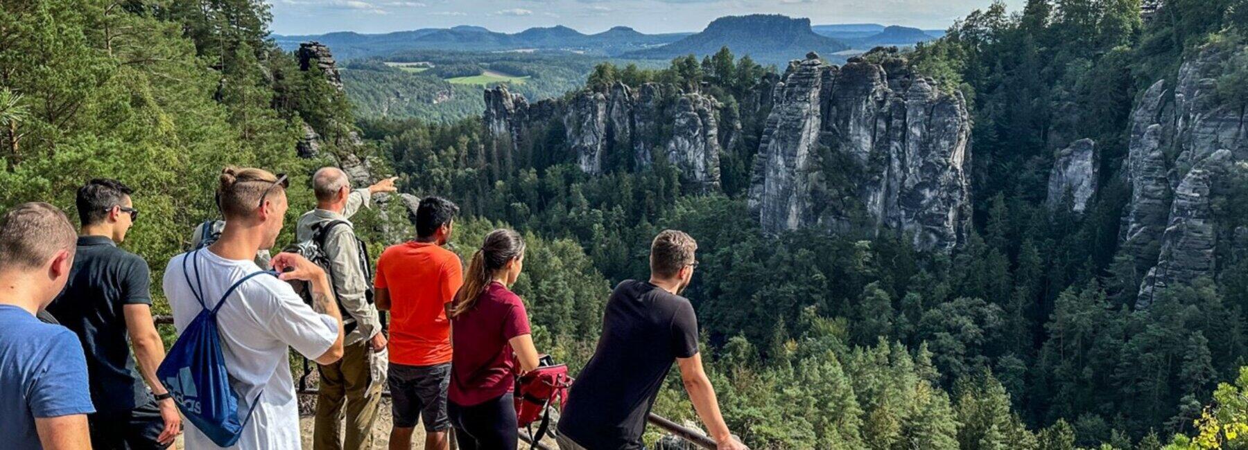 A group of hikers is standing on a plateau, looking at sandstone cliffs