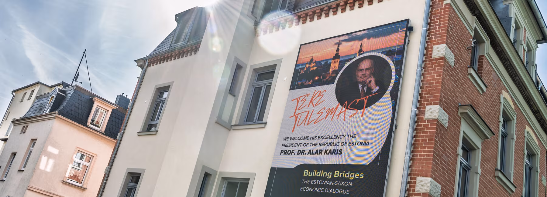Eine Hausfassade mit einem wandfüllenden LED-Display: Es zeigt das Foto von Alar Karis und unter anderem den Text „Building Bridges“. Im gegen die Sonne gemachten Foto spiegeln sich die Linsen des Objektivs.