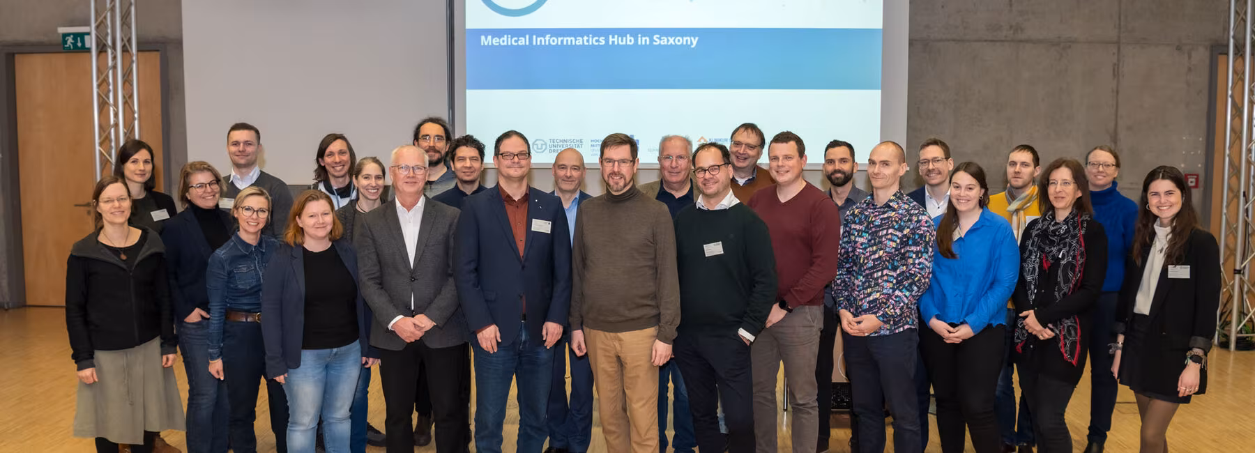 Gruppenaufnahme mit 26 Personen. In Hintergrund ist eine Folie projiziert mit dem MiHUBx-Logo und dem Schriftzug „Medical Informatics Hub in Saxony“.