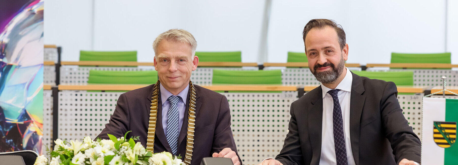 Zwei männliche Personen mit Anzug und Krawatte sitzen an Abgeordnetenbänken eines Plenarsaals. Die linke Person trägt eine goldene Amtskette. Vor ihnen liegen aufgeschlagene Dokumente, die auf der linken Seite von einem Blumengesteck zum größten Teil verdeckt werden. Am rechten Bildrand ist ein Tischbanner mit dem sächsischen Wappen zu sehen.