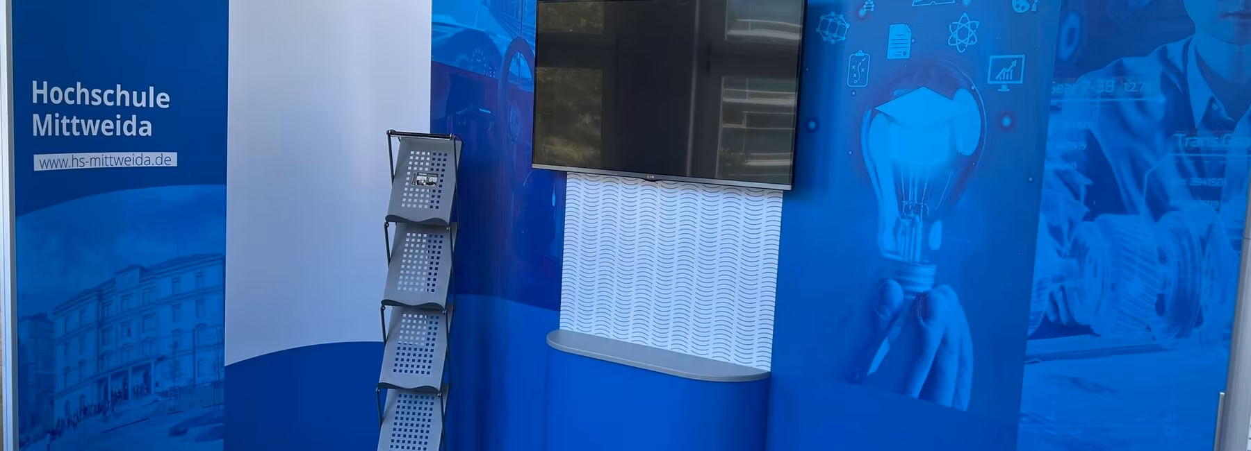 Elemente eines messesatndes, ein Bildschirm an der Wand, ein Flyerständer vor dem Stand