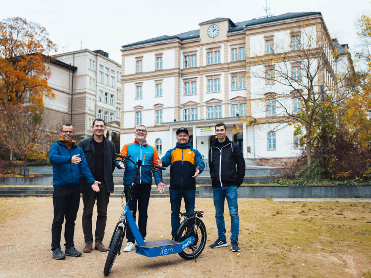 Zwei Hochschulgebäude im Hintergrund, im Vordergrund: fünf männliche Personen stehen um einen E-Scooter