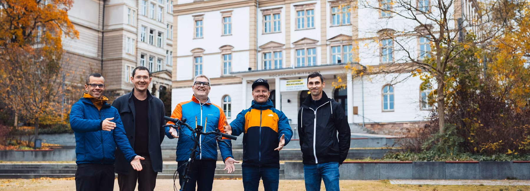 Two university buildings in the background; in the foreground: five men are standing around an e-scooter