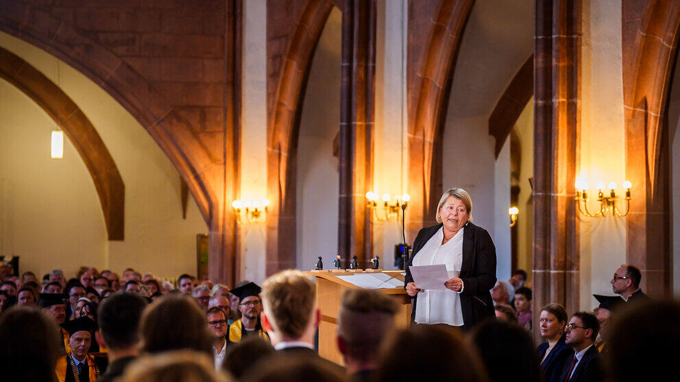 Festrednerin Dr. Sabine Hückmann wendet sich vom Rednerpult aus nach hinten zu den Absolvent:innen.