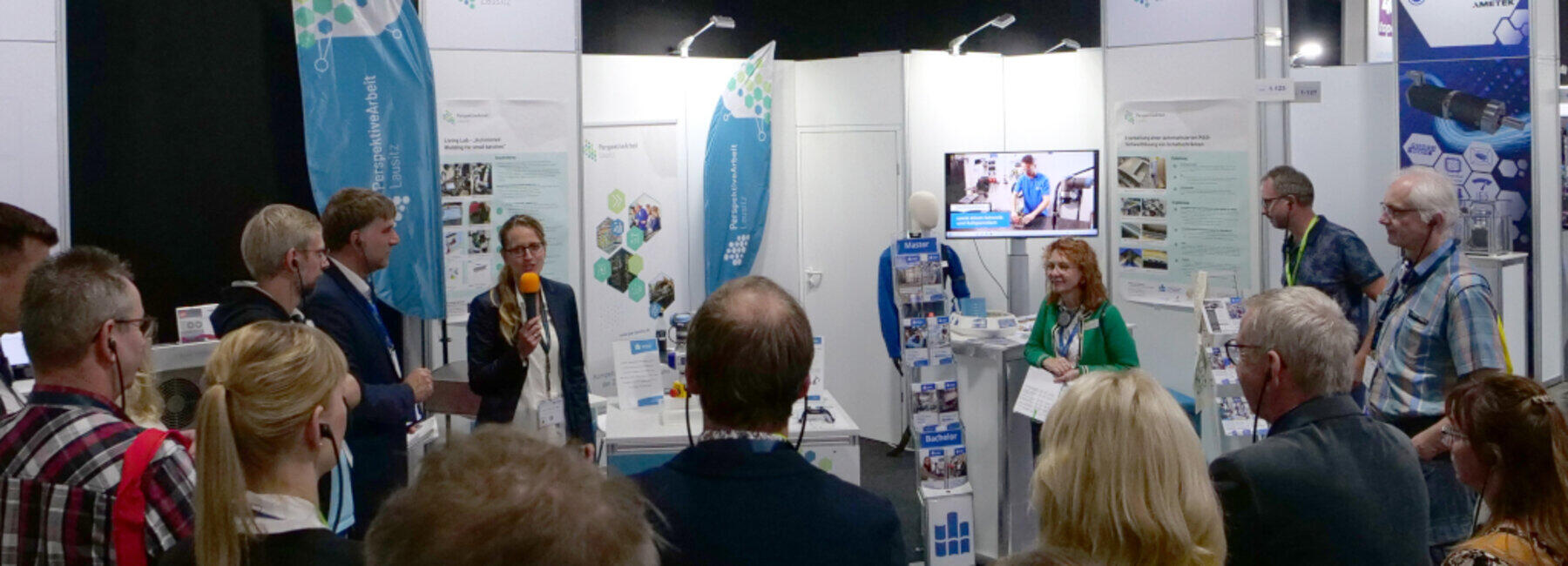 Exhibition stand; people are standing in a semicircle; one person is speaking into a microphone
