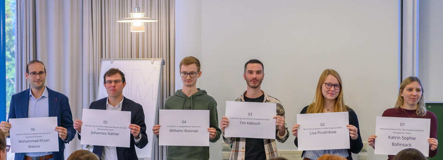 Six people are each holding up a large name badge in front of an audience