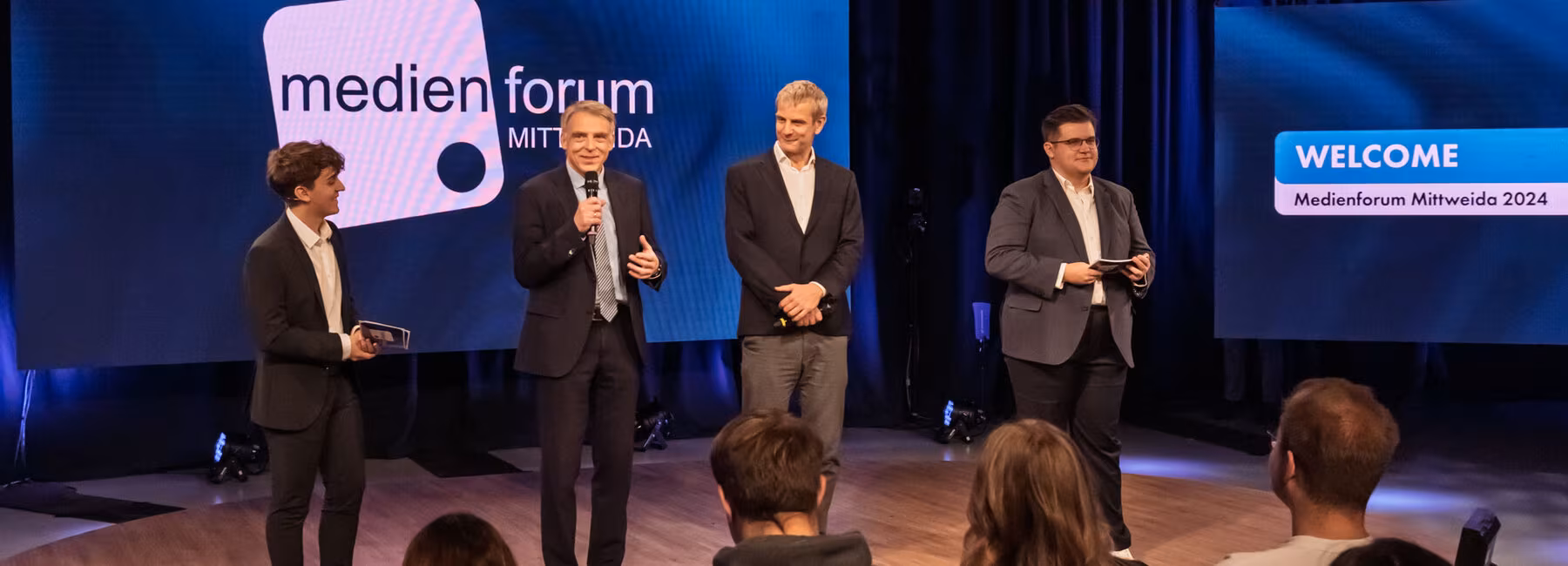 Vier männliche Personen stehen auf einem leicht erhöhten Podest im Fernsehstudio. Im Vordegrund sitzen Zuschauer:innen. Auf den Screens im Hintergrund sind das Medienforum-Mittweida-Logo und der Schriftzug "WELCOME. Medienforum Mittweida 2024" zu sehen..