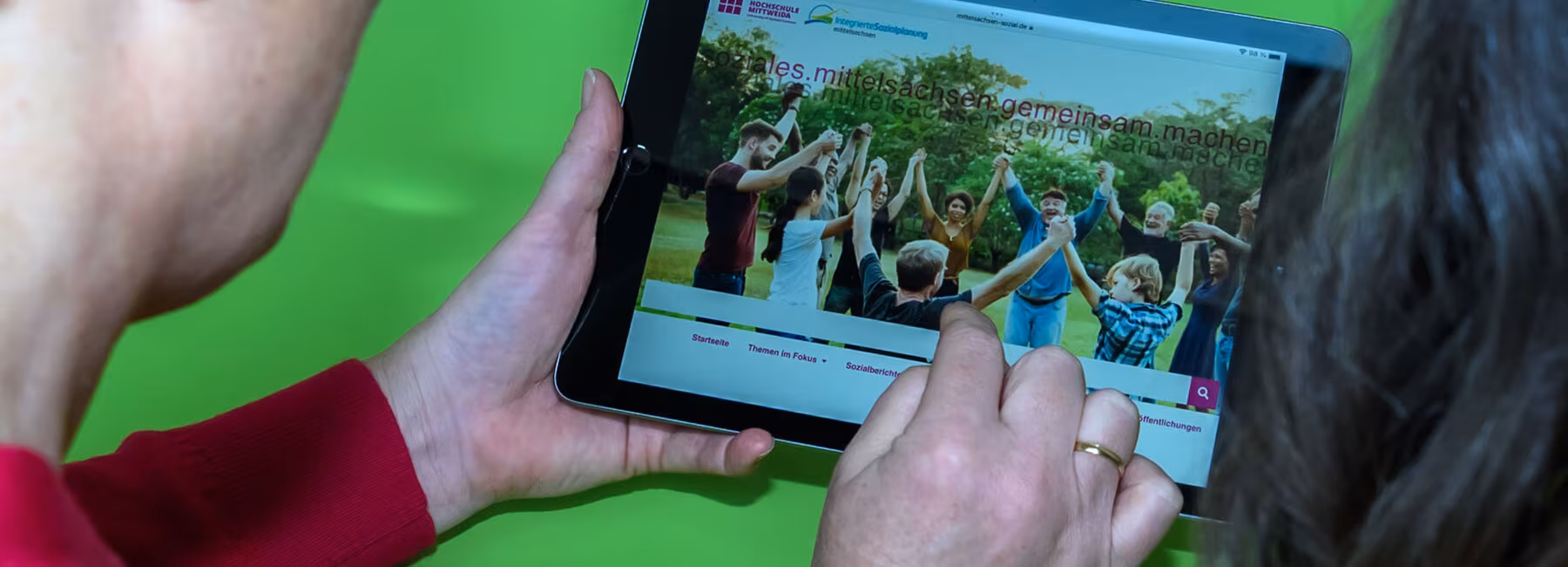 Blick über die Schultern zweier Personen auf ein Tablet in den Händen der linken Person. Sie zeigt mit einem Finger der rechten Hand auf die Oberfläche des Tablets. Es zeigt eine Webseite mit einem Bildmotiv mit Menschen unterschiedlichen Geschlechts und Alters, die im Kreis stehend sich an den Händen fassen. Über das Bildmotiv ist zweimal der Schriftzug mit unterschiedlicher Deckkraft „soziales.mittelsachsen.gemeinsam.machen“ gelegt.