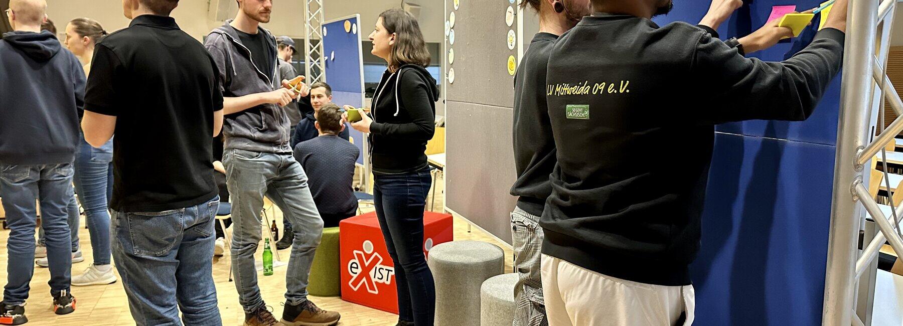 Gruppe von Personen steht im Raum und vor einer Pinnwand