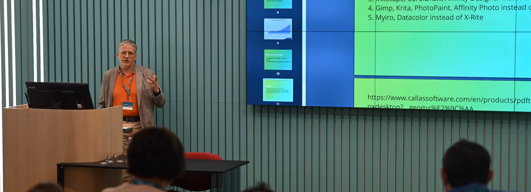 Referent steht im Hörsaal vor Anwesenden und im Hintergrund ist seine Präsentation auf der Leinwand zu sehen