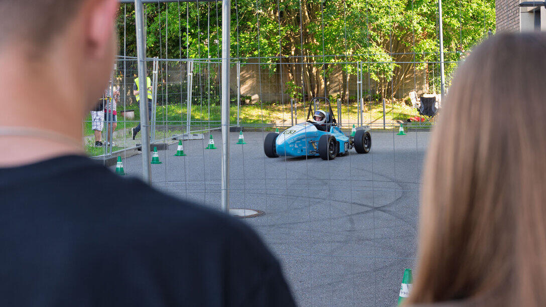 Blick zwischen die angeschnittenen Köpfe zwier Zuschauender auf einen Formula-Student-Rennwagen, der auf inem mit Bauzäunen abgesperrten Parcours fährt.