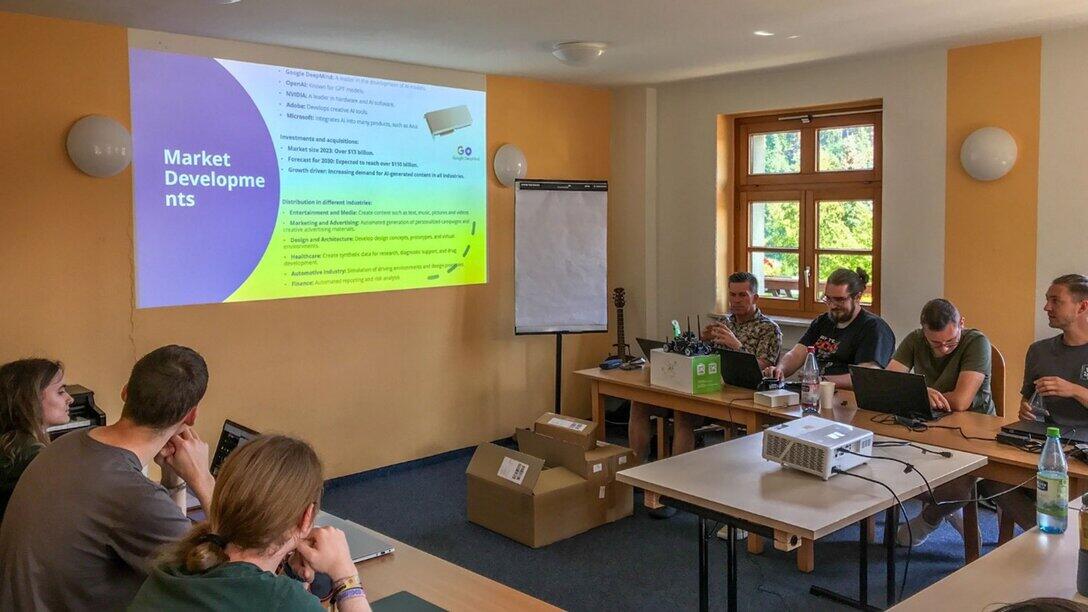 Gruppe von personen sitzt in einem Raum an Tischen und folgt einer Präsentation