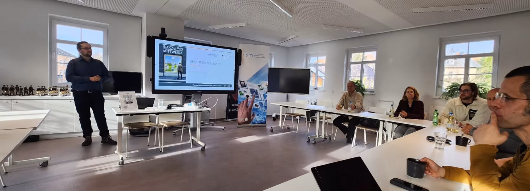 Männliche person steht vor Präsentationswand, weitere Personen sitzen im Raum