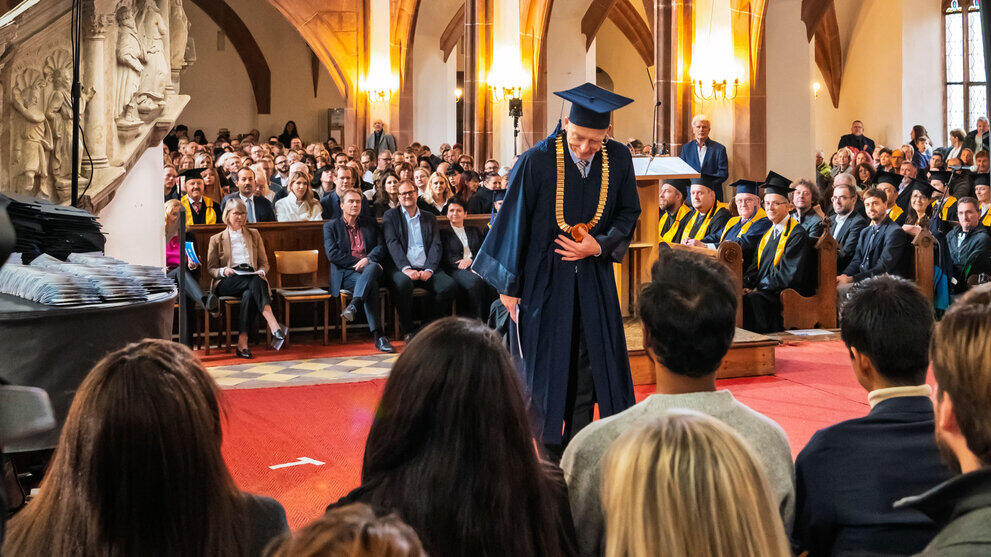 Männliche Person im blauen Talar, mit goldener Amtskette udn Doktorhut verneigt sich vor sitzenden Personen im Bildvordergrund.