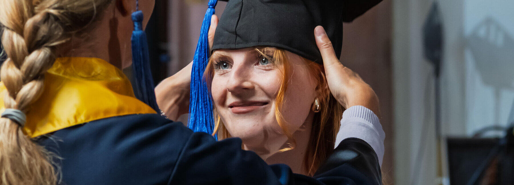 Weibliche Person im Talar und mit Doktorhut setzt Absolventin den Graduierten-Hut auf.