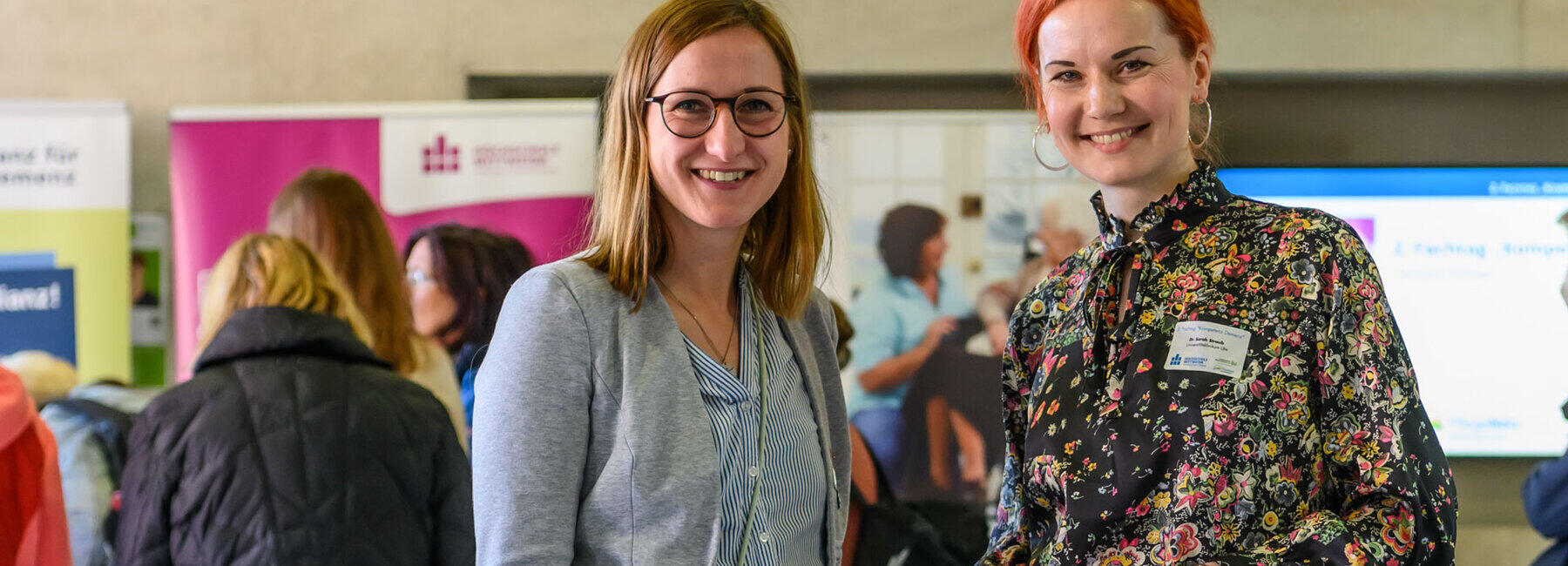 Zwei jüngere Frauen stehen in einem Foyer nebeneinander und schauen strahlend in die Kamera. Im Hintergrund sind unscharf weitere Menschen vor Rollups zu sehen. Auf einem der Rollups ist das Logo der Hochschule Mittweida zu erkennen.