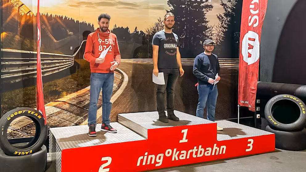 Drei junge männliche Personen stehen auf einem dreistufigen Siegerpodest und halten Urkunden in den Händen. Auf dem Podest ist weiß auf rot „Ring Kartbahn“ zu lesen. Rennreifen sind links und rechts vom Podest drapiert. Der Hintergrund ist die Abbildung eines Ausschnitts einer Rennkurve mit Leitplanken.