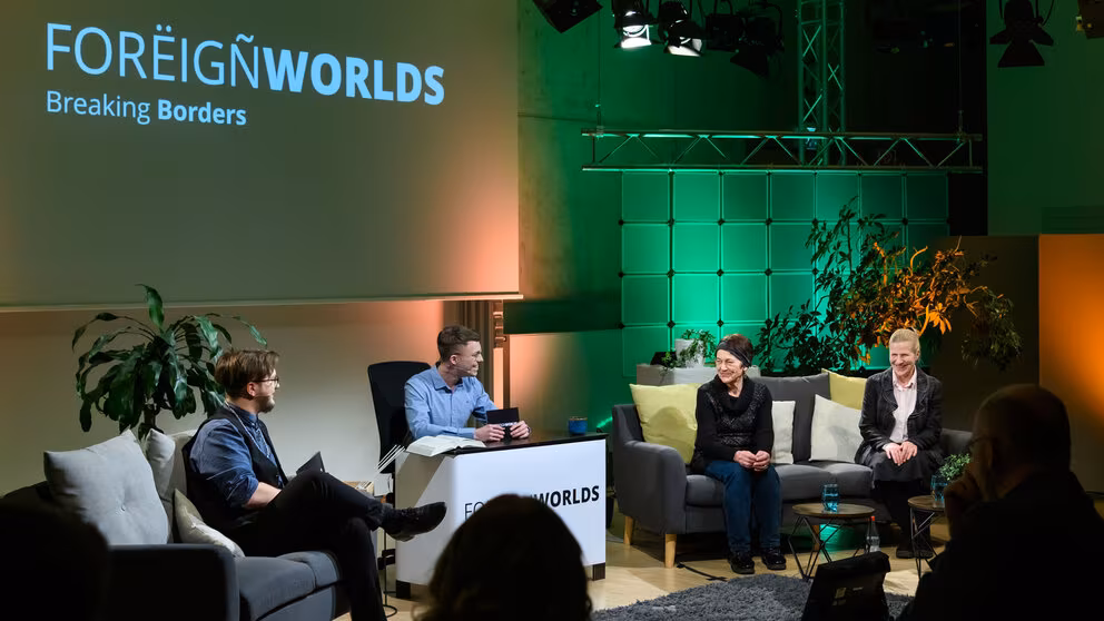 Die beiden Moderatoren wenden ihren Blick zwei weiblichen Personen auf einem Sofa rechts im Bild. Die Stimmung ist heiter.