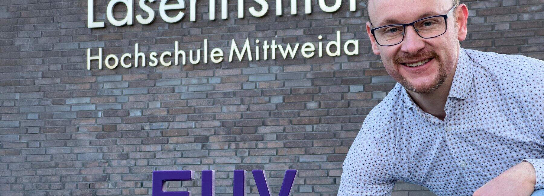 Männliche Person hockt vor Hauswand mit der Aufschrift Laserinstistut Hochschule Mittweida, neben ihm steht ein Schriftzug EUV ein