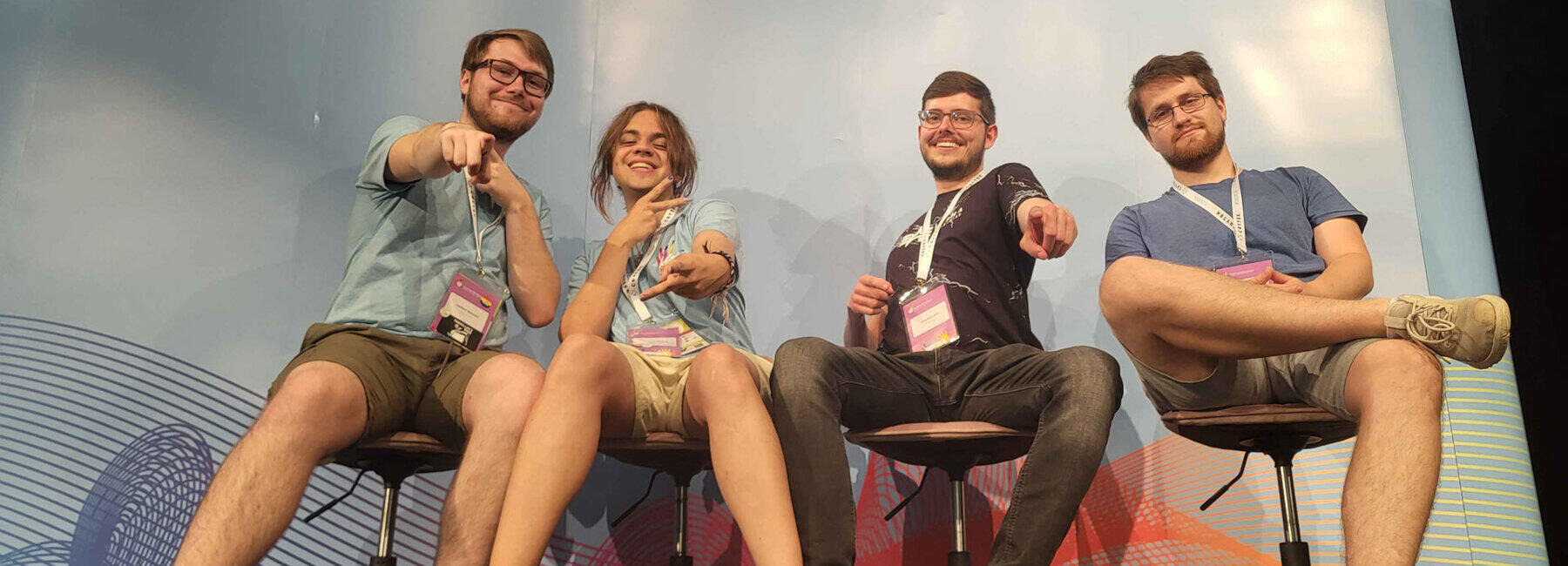 Vier junge Menschen sitzen fröhlich gestikulierend vor eine Messewand mit dem Schriftzug „German DevDays. INDIE AWARD“.